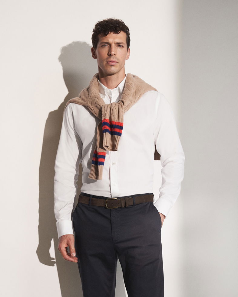 Mann mit weißem Hemd, dunkelblauer Hose, braunem Gürtel und beigem Pullover mit roten und blauen Streifen um die Schultern.