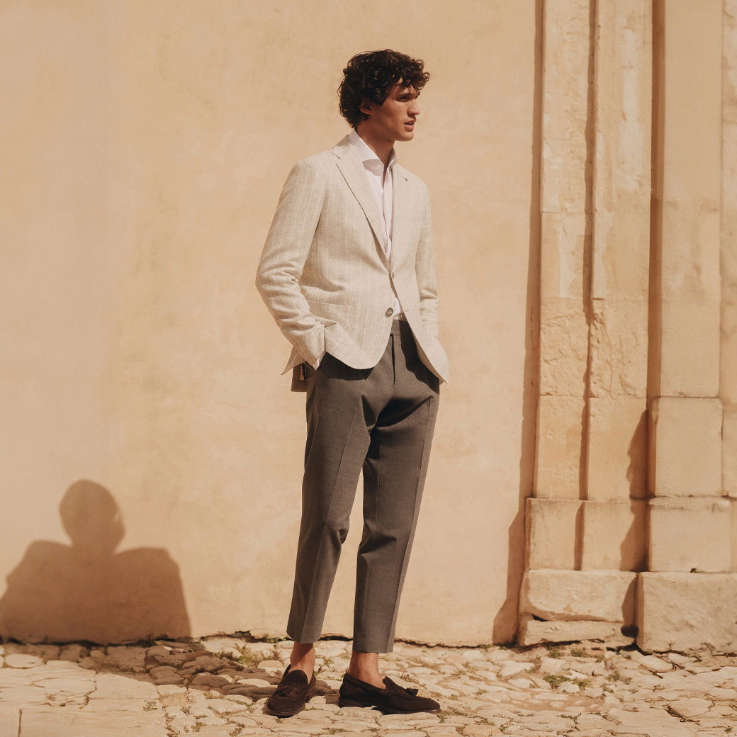 Mann steht vor Wand, trägt beige gestreiftes Sakko, weißes Hemd, braune Hose und braune Loafer ohne Socken. Mann steht vor Wand, trägt beige gestreiftes Sakko, weißes Hemd, braune Hose und braune Loafer ohne Socken.