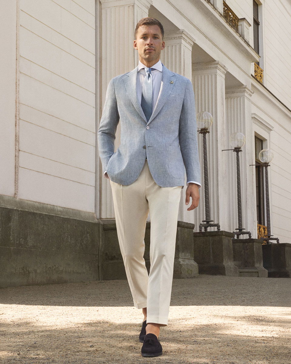 Mann in cremefarbener Hose, hellblauem Sakko, weißem Hemd, blauer Krawatte und dunkelblauen Loafern vor weißem Gebäude.