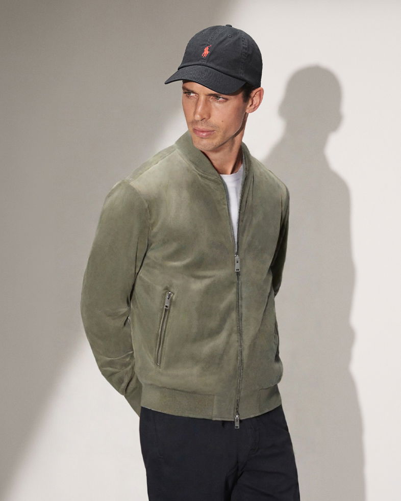 Mann trägt olivgrünen Veloursleder-Blouson, weißes Shirt, dunkelblaue Hose und schwarze Cap, schaut seitlich weg.