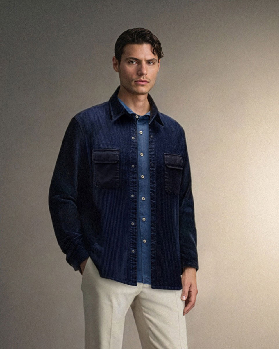 Mann mit dunkelblauem Overshirt, blauem Jeanshemd und beige Hose, vor neutralem Hintergrund stehend.