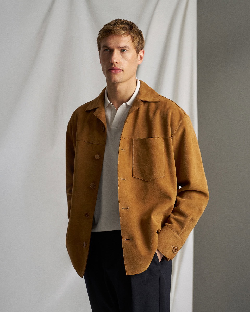 Mann im Studio trägt hellbraune Velourslederjacke, weißes Poloshirt und navyblaue Hose, steht vor hellem Hintergrund.