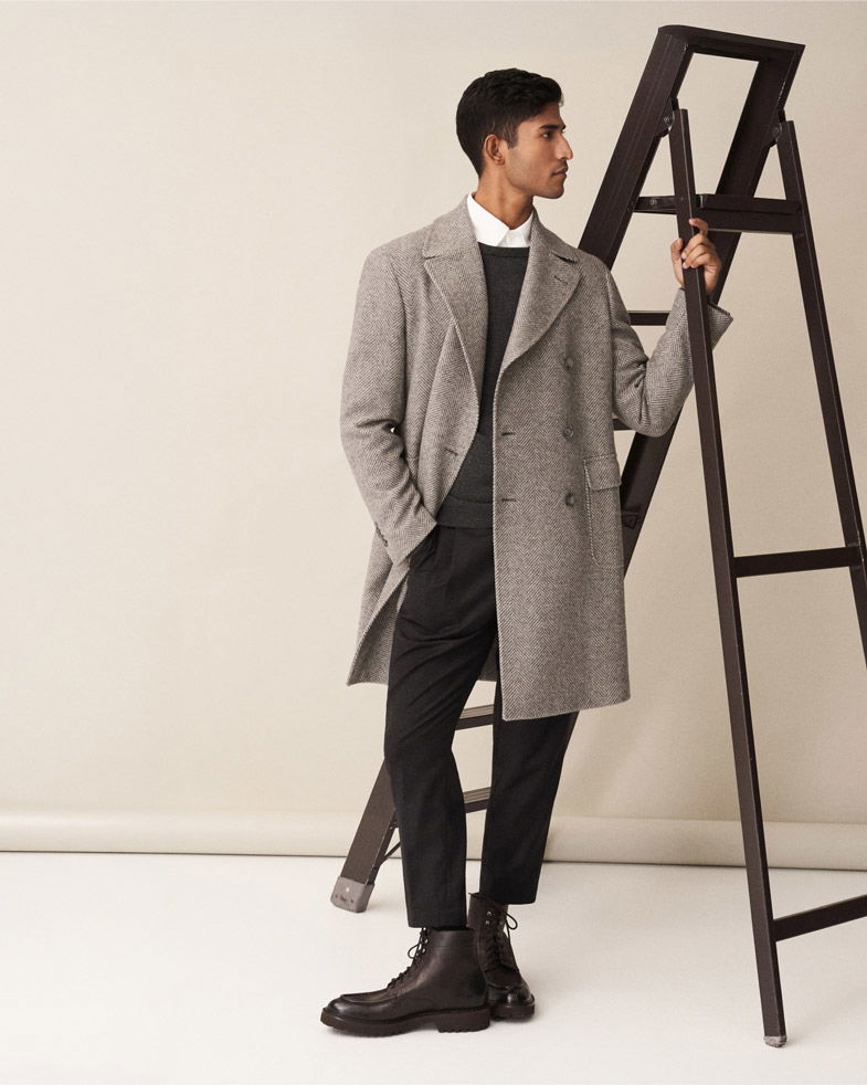 Mann lehnt an brauner Leiter, trägt hellgrauen Mantel, schwarzen Pullover, weißes Hemd, schwarze Hose und braune Schnürboots.