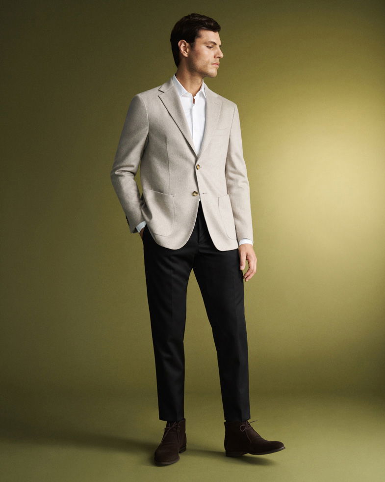 Mann trägt beigen Sakko, weißes Hemd, schwarze Hose und dunkelbraune Desert Boots, steht vor grünem Hintergrund.