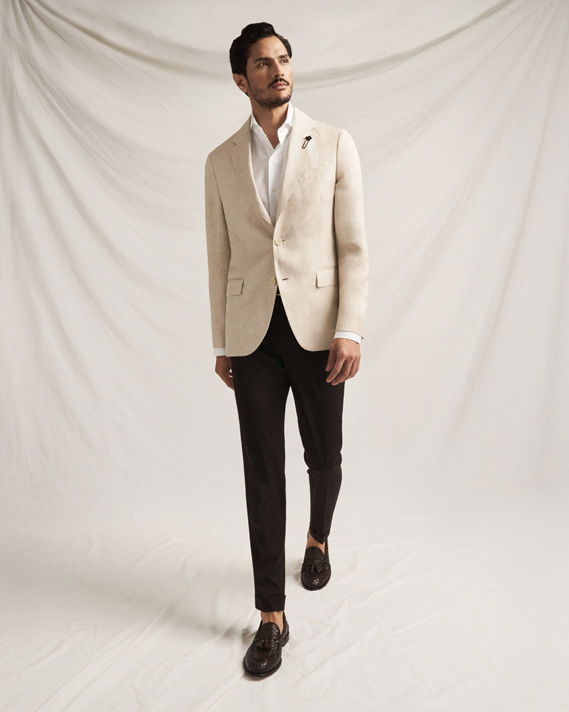 Mann in beige Sakko, weißem Hemd, schwarzer Hose und dunkelbraunen Loafern vor hellem Stoffhintergrund, stehend und nach rechts schauend. Mann in beige Sakko, weißem Hemd, schwarzer Hose und dunkelbraunen Loafern vor hellem Stoffhintergrund, stehend und nach rechts schauend.