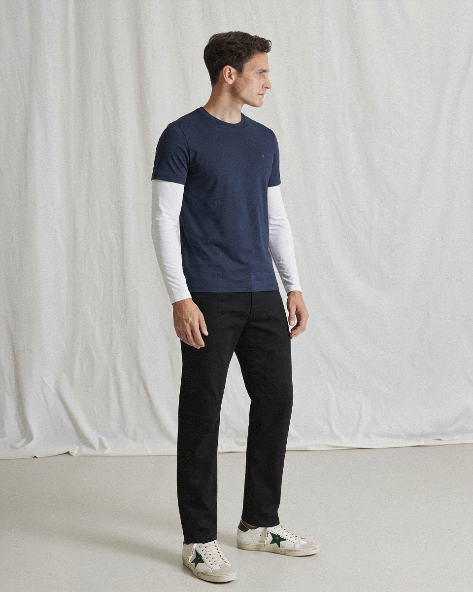 Mann in dunkelblauem T-Shirt über weißem Langarmshirt, schwarzer Hose und weißen Sneakers mit grünem Stern vor hellem Hintergrund.