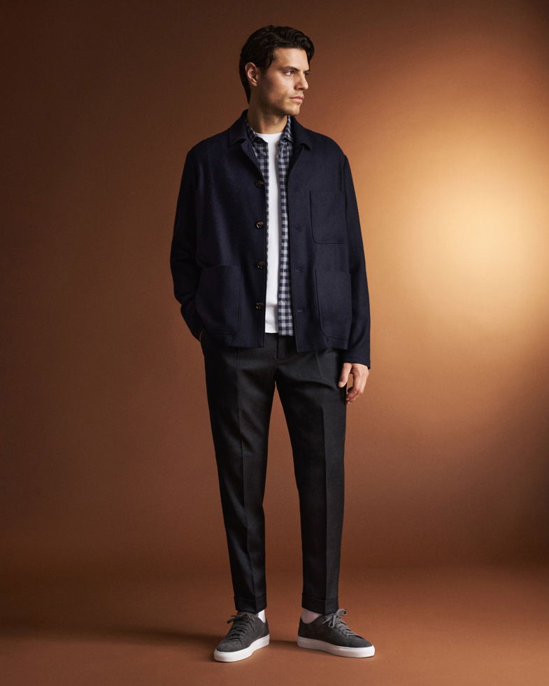 Mann in dunkelblauem Overshirt, weißem T-Shirt, kariertem Hemd, schwarzer Hose und grauen Sneakern vor braunem Hintergrund. Mann in dunkelblauem Overshirt, weißem T-Shirt, kariertem Hemd, schwarzer Hose und grauen Sneakern vor braunem Hintergrund.