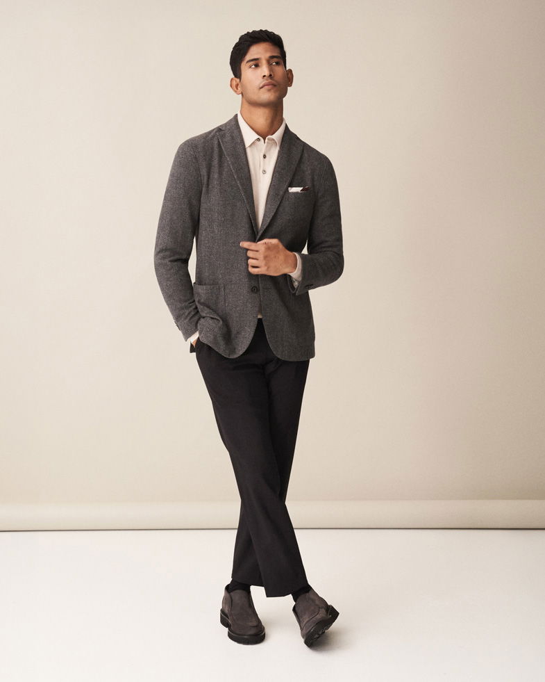 Mann trägt dunkelgraues Sakko, cremefarbenes Strickpolo, schwarze Hose und dunkelgraue Chukka-Boots vor hellem Hintergrund.