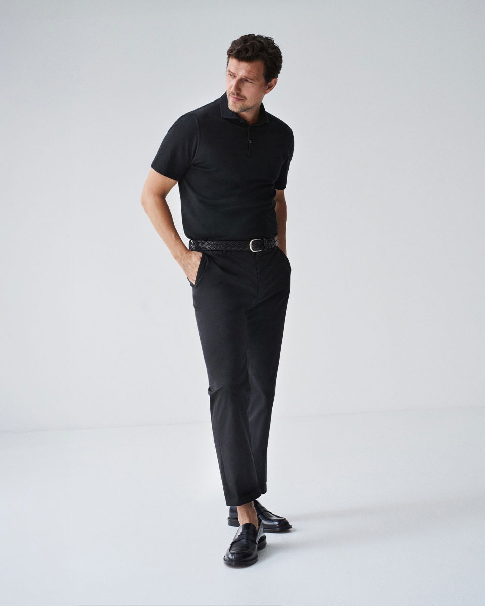 Mann trägt schwarzes Poloshirt, schwarze Hose, schwarzen geflochtenen Ledergürtel und schwarze Lederloafer, steht vor weißem Hintergrund.