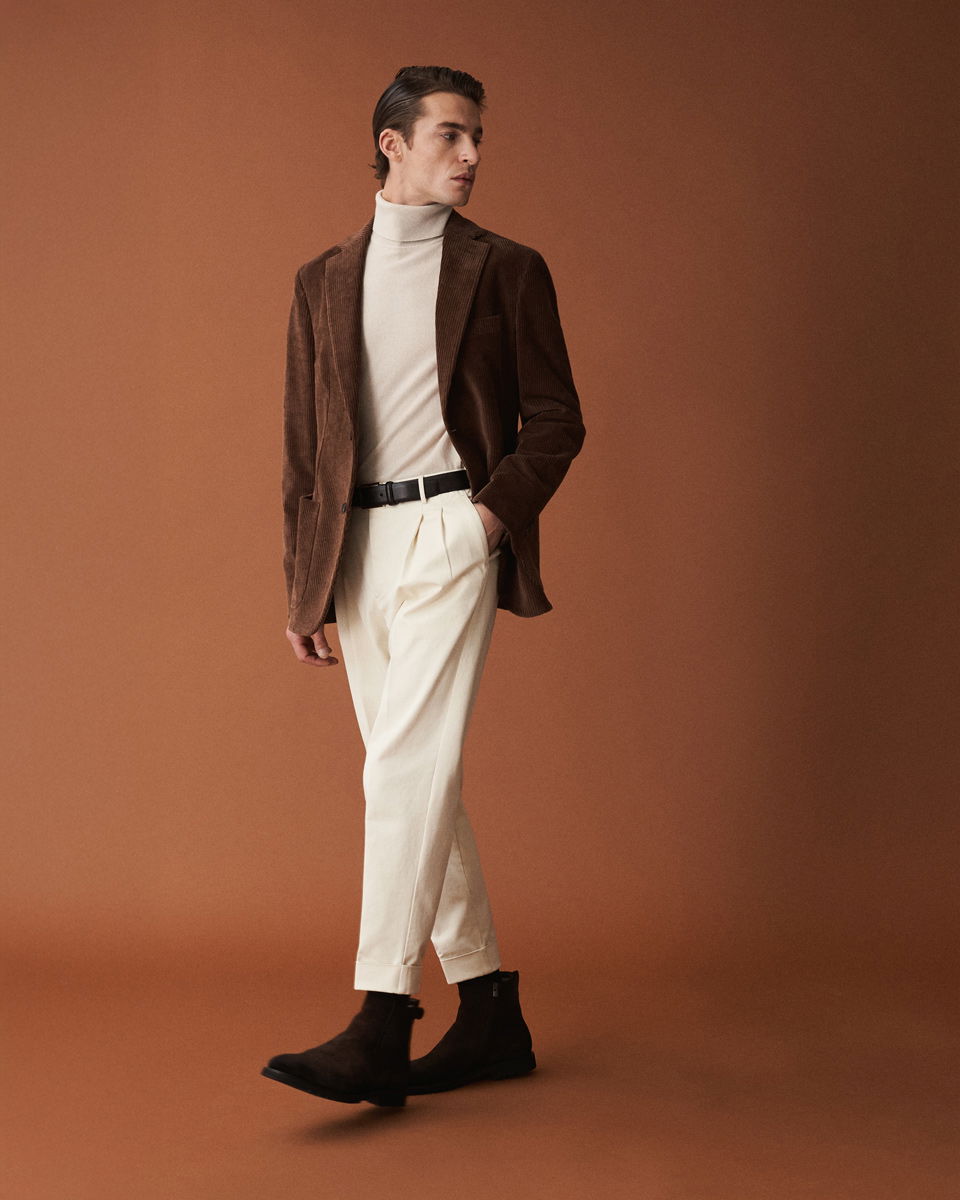 Herr trägt beige Rollkragenpullover, braunes Cord-Sakko, weiße Hose, schwarzen Gürtel und braune Chelsea Boots, brauner Hintergrund.
