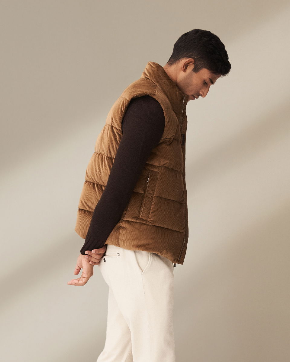 Mann in hellbrauner Cord-Daunenweste, dunkelbraunem Pullover und weißer Hose, seitlich stehend, Hände hinter dem Rücken.