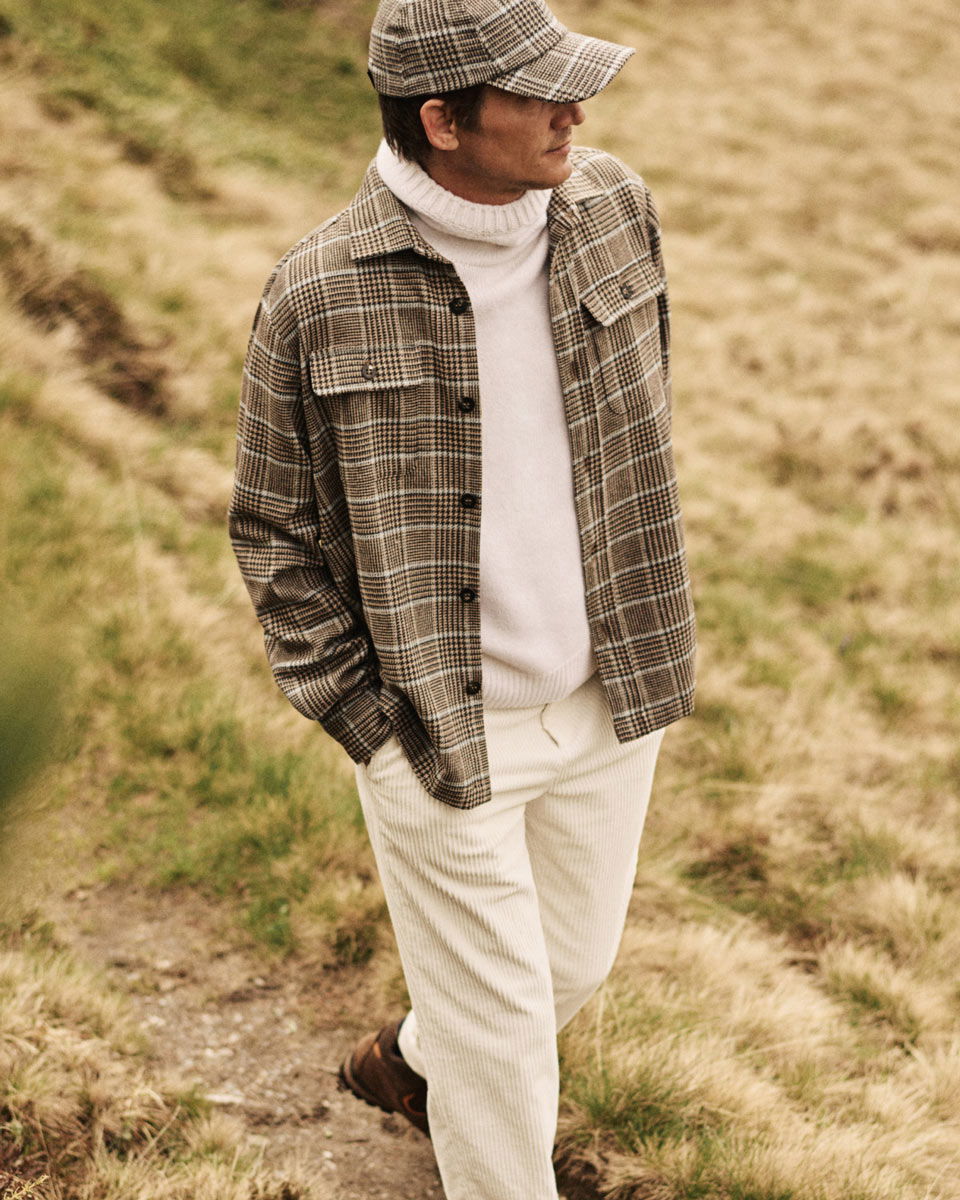 Mann trägt braun-weiß kariertes Overshirt, weiße Cordhose, weißen Rollkragenpullover und kariertes Cap, steht im Gras. Mann trägt braun-weiß kariertes Overshirt, weiße Cordhose, weißen Rollkragenpullover und kariertes Cap, steht im Gras.
