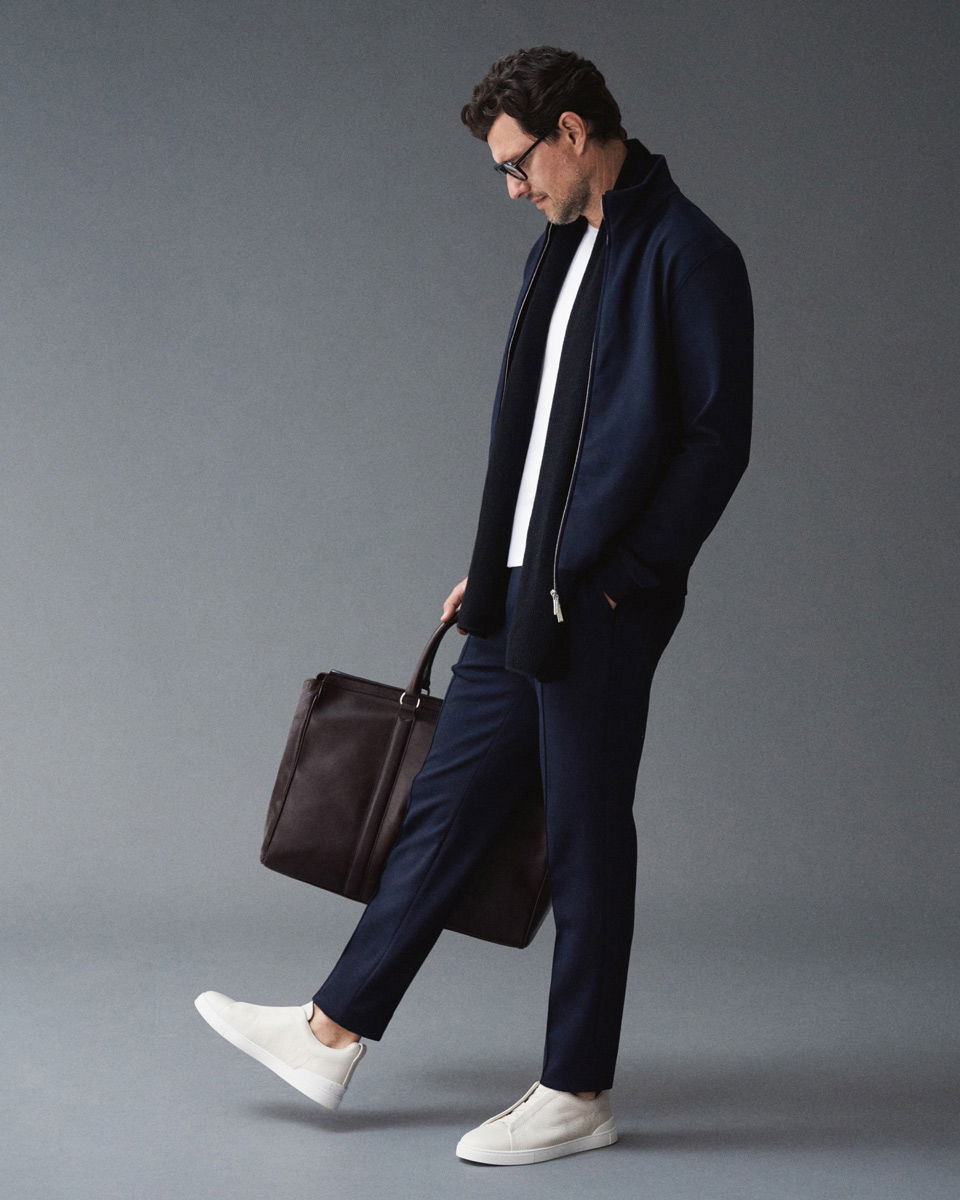 Mann in navyblauer Jacke und Hose, weißem T-Shirt, schwarzem Schal, cremefarbenen Sneakern, trägt braunen Weekender vor grauem Hintergrund.