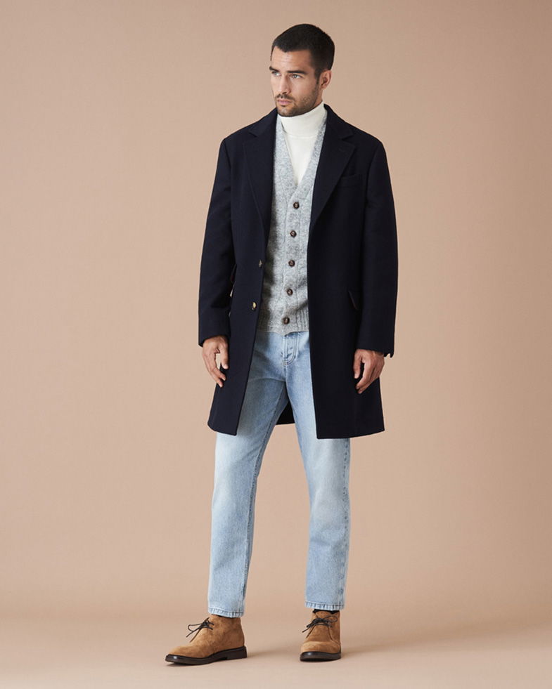 Mann trägt hellblauen Jeans, weißen Rollkragenpullover, hellgrauen Cardigan, navyblauen Mantel und hellbraune Desert Boots.