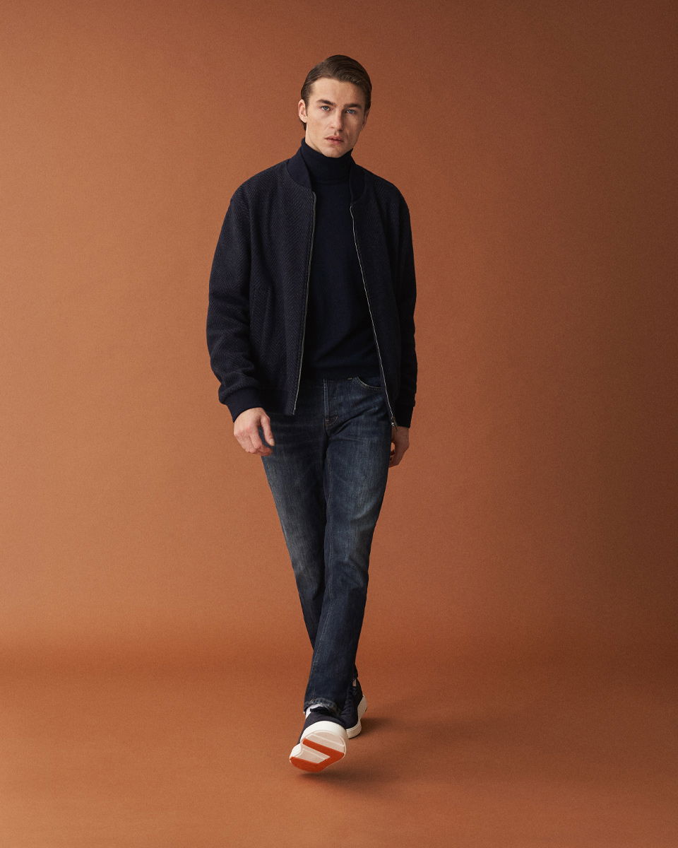 Mann geht vor braunem Hintergrund, trägt dunkle Jeans, schwarzen Rollkragenpullover, dunkelblauen Blouson und schwarze Sneaker.