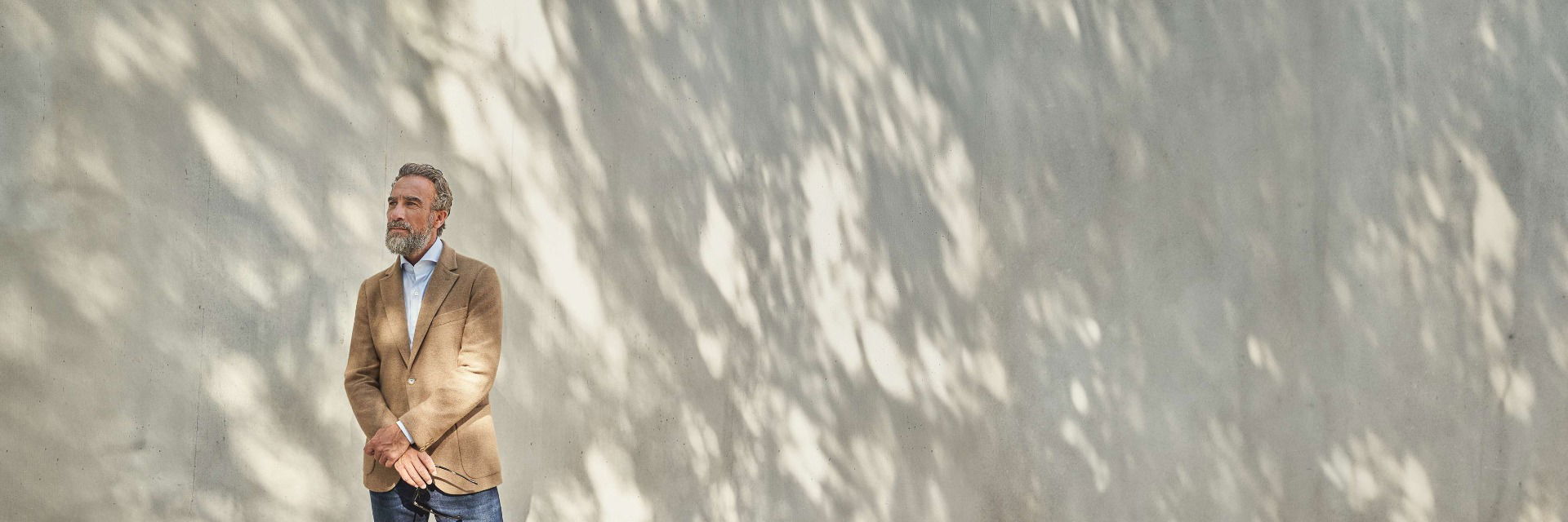 Mann mit grauem Bart trägt hellbraunes Sakko, hellblaues Hemd und Jeans, steht vor sonniger Betonwand mit Schatten. Mann mit grauem Bart trägt hellbraunes Sakko, hellblaues Hemd und Jeans, steht vor sonniger Betonwand mit Schatten.