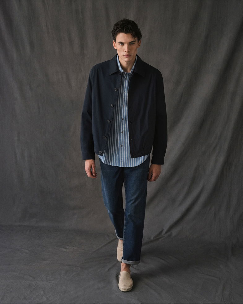 Junger Mann in dunkelblauem Overshirt, blau-weiß gestreiftem Hemd, blauen Jeans und beigen Espadrilles vor grauem Hintergrund.