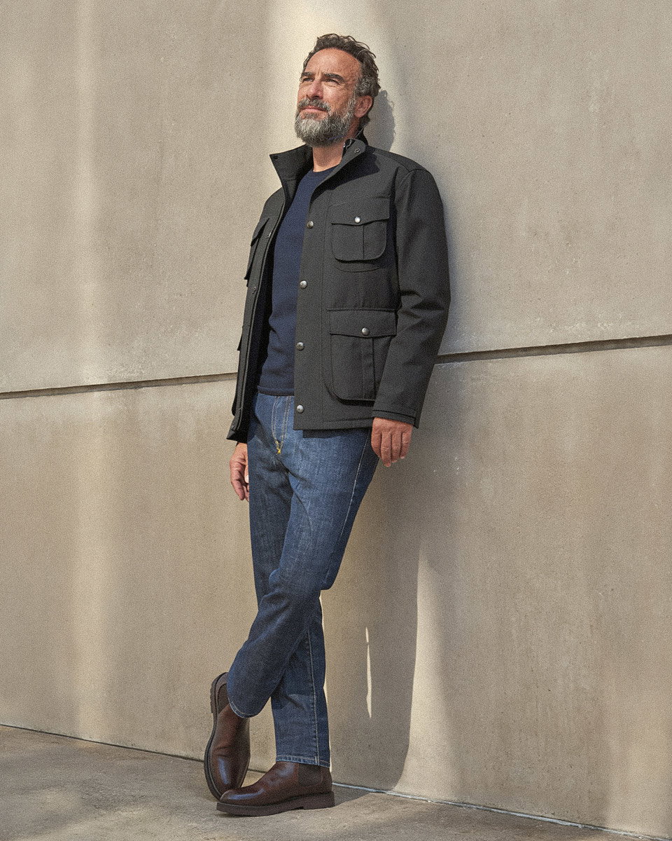 Mann mit grau meliertem Bart lehnt an Wand, trägt schwarze Jacke, dunkelblaues Shirt, blaue Jeans und braune Chelsea Boots.