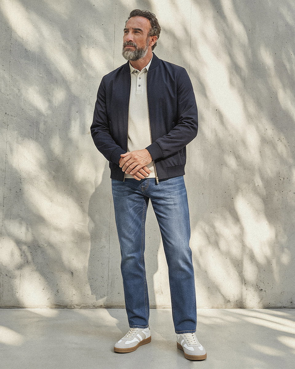 Mann mit blau-grauem Poloshirt, dunkelblauem Blouson, blauer Jeans und weiß-grauen Sneakern vor heller Betonwand.