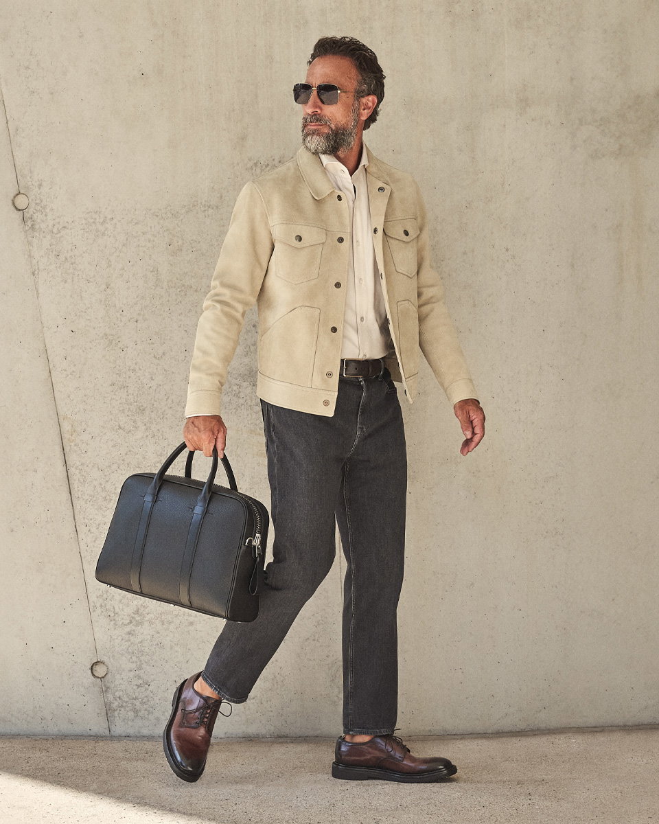 Mann mit beiger Jacke, weißem Hemd, dunkelgrauer Jeans, braunen Schnürschuhen, schwarzer Tasche und Sonnenbrille vor Betonwand.