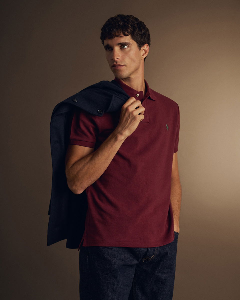 Mann trägt dunkelrotes Poloshirt, hält dunkelblaues Overshirt über Schulter, dazu dunkle Jeans, Blick seitlich, brauner Hintergrund. Mann trägt dunkelrotes Poloshirt, hält dunkelblaues Overshirt über Schulter, dazu dunkle Jeans, Blick seitlich, brauner Hintergrund.