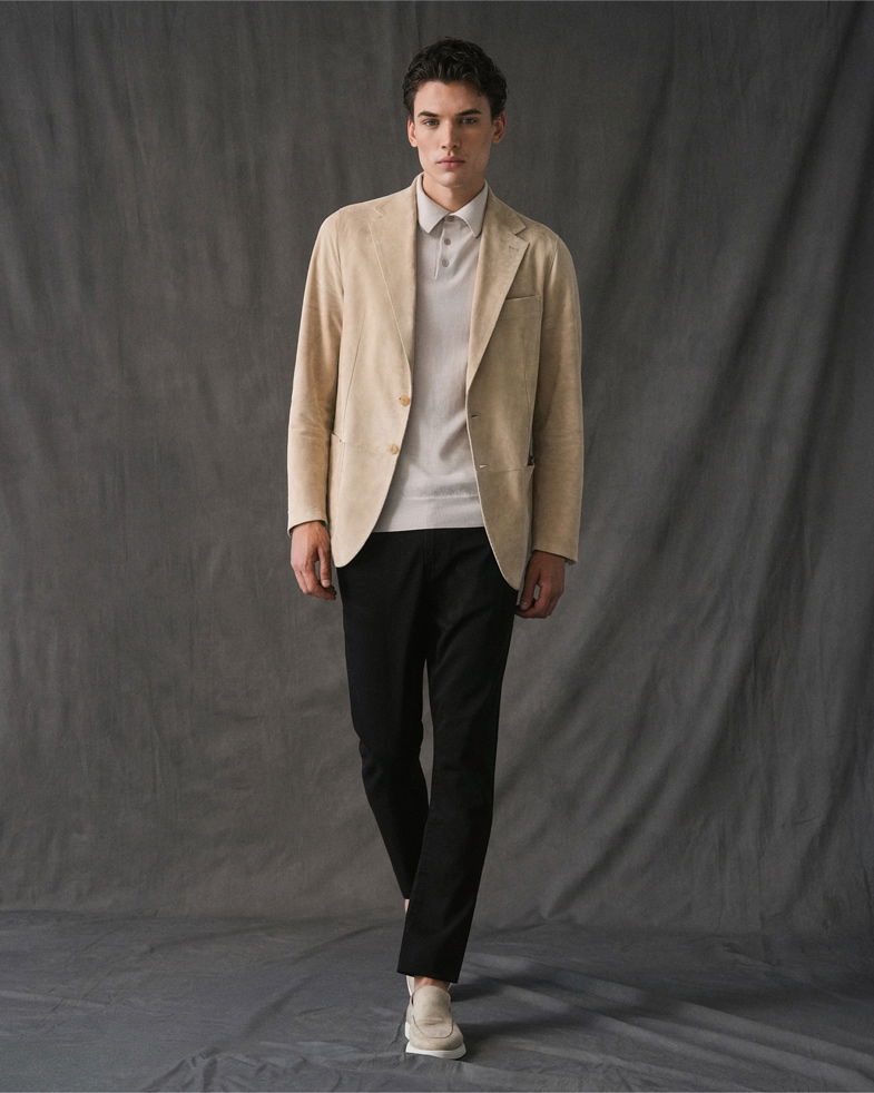 Junger Mann mit beigem Sakko, cremefarbenem Polohemd, schwarzer Hose und hellen Loafers vor grauem Hintergrund.