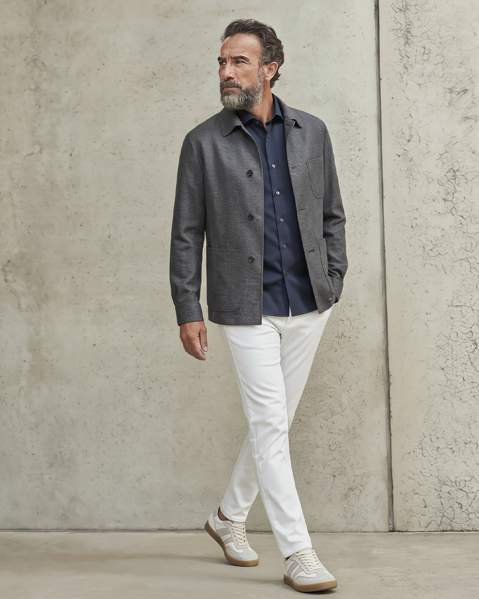 Mann mit grauem Overshirt, dunkelblauem Hemd, weißen Hosen und weißen Sneakern vor heller Betonwand, nach links schauend.
