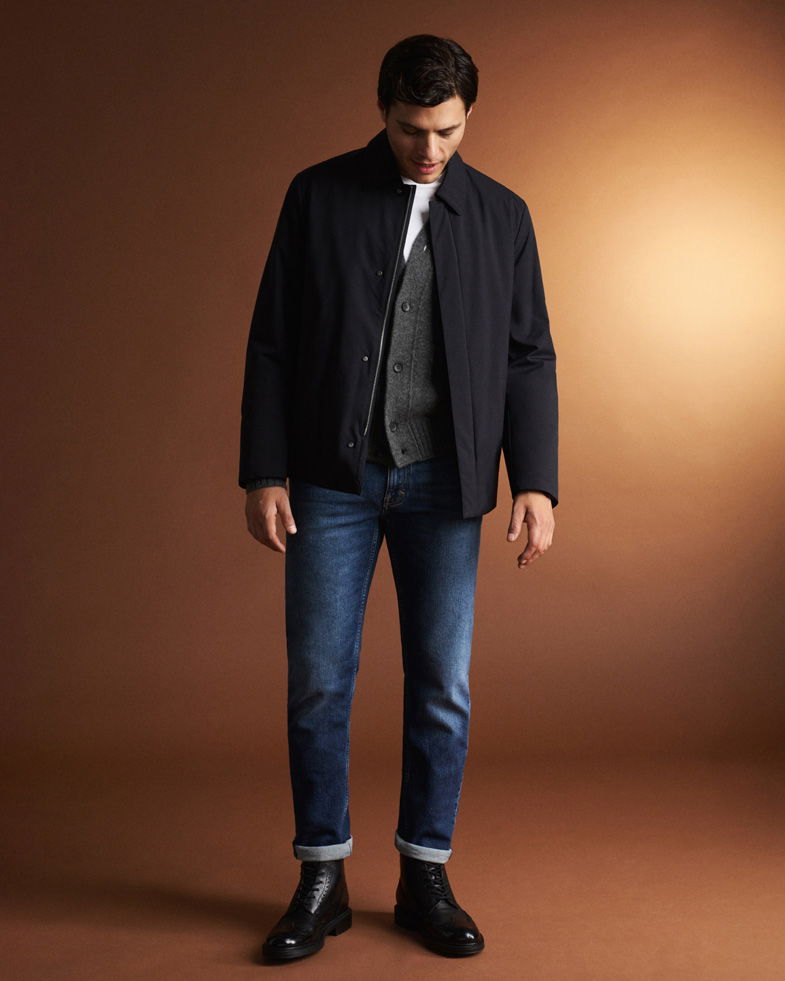 Mann trägt weiße Shirt, dunkelgrauen Cardigan, navy Jacke, blaue Jeans und schwarze Boots vor braunem Hintergrund. Mann trägt weiße Shirt, dunkelgrauen Cardigan, navy Jacke, blaue Jeans und schwarze Boots vor braunem Hintergrund.
