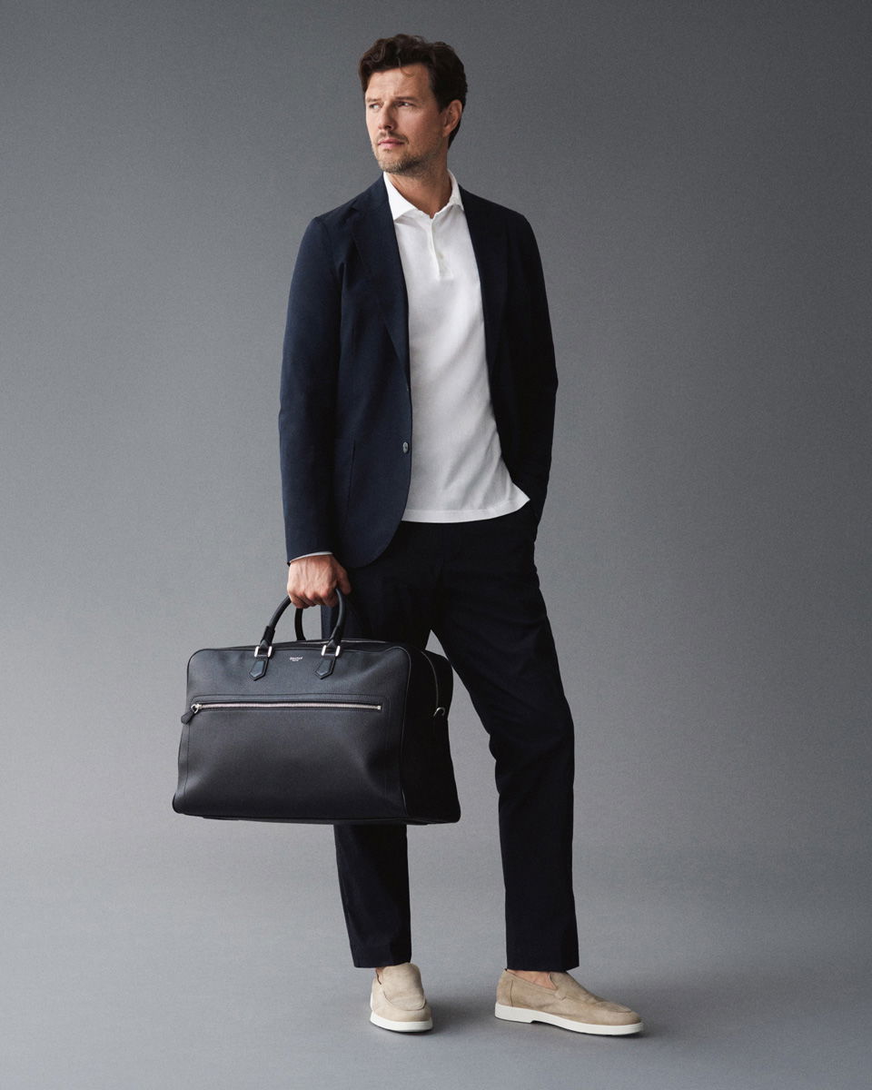 Mann trägt navy Jersey-Sakko und Hose, weißes Langarm-Poloshirt, beige Loafer, hält schwarzen Weekender in der Hand.