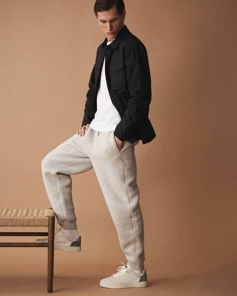 Mann trägt hellgraue Jogginghose, weißes T-Shirt, schwarzes Overshirt und weiße Sneaker, steht mit einem Fuß auf einer Bank.