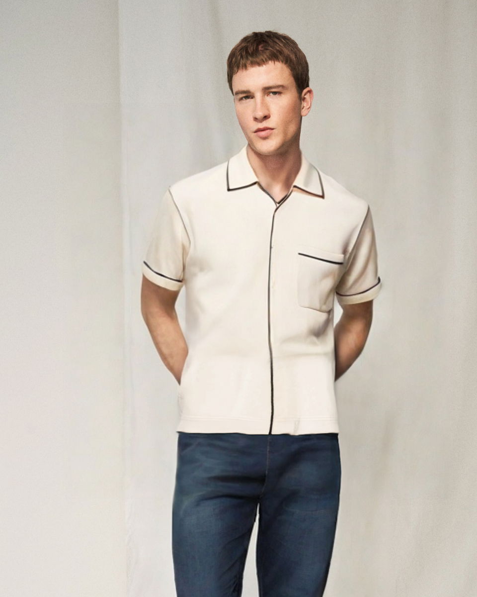 Mann mit kurzem braunen Haar trägt cremefarbenes Kurzarmhemd mit dunklen Saumkanten und dunkelblaue Jeans vor hellem Hintergrund. Mann mit kurzem braunen Haar trägt cremefarbenes Kurzarmhemd mit dunklen Saumkanten und dunkelblaue Jeans vor hellem Hintergrund.