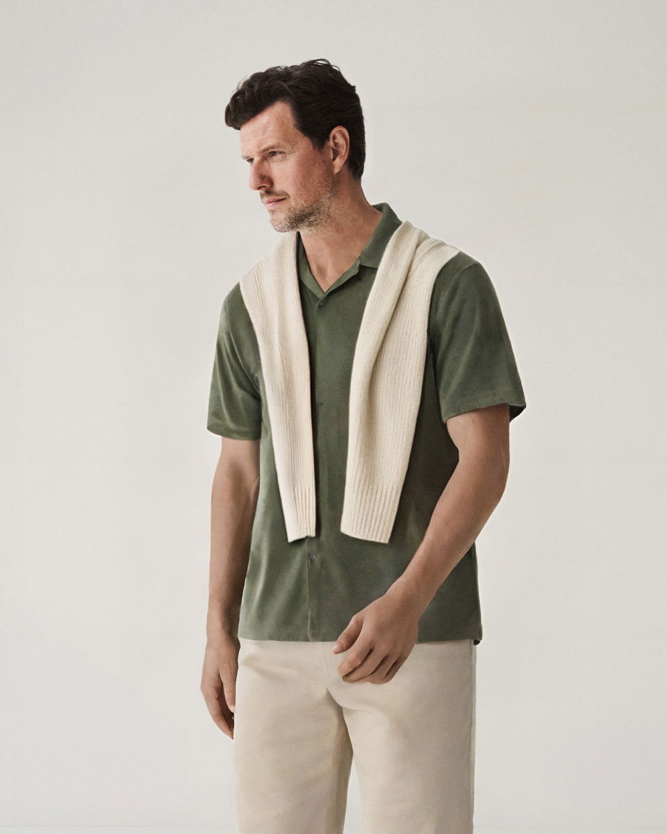 Mann mit olivgrünem Kurzarmhemd, beiger Hose und beigem Pullover locker über die Schultern gelegt, vor hellem Hintergrund.