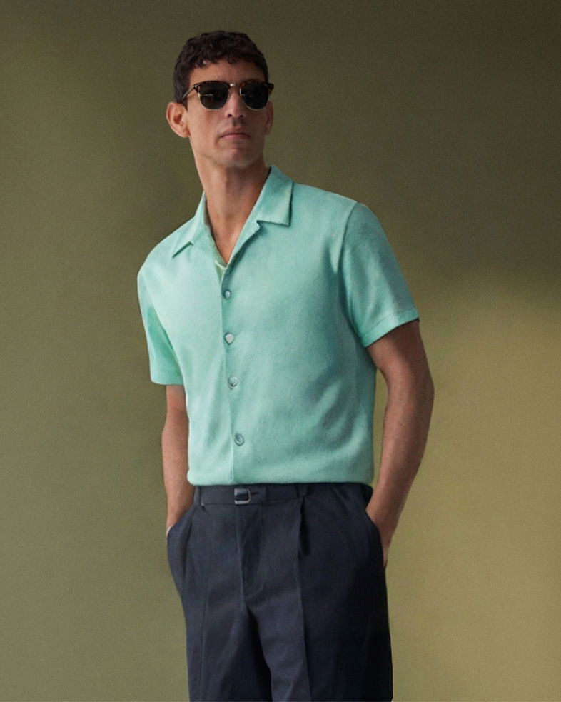 Mann mit kurzärmligem türkisfarbenem Hemd, navy Hose, Sonnenbrille, Hände in Taschen vor grünem Hintergrund.