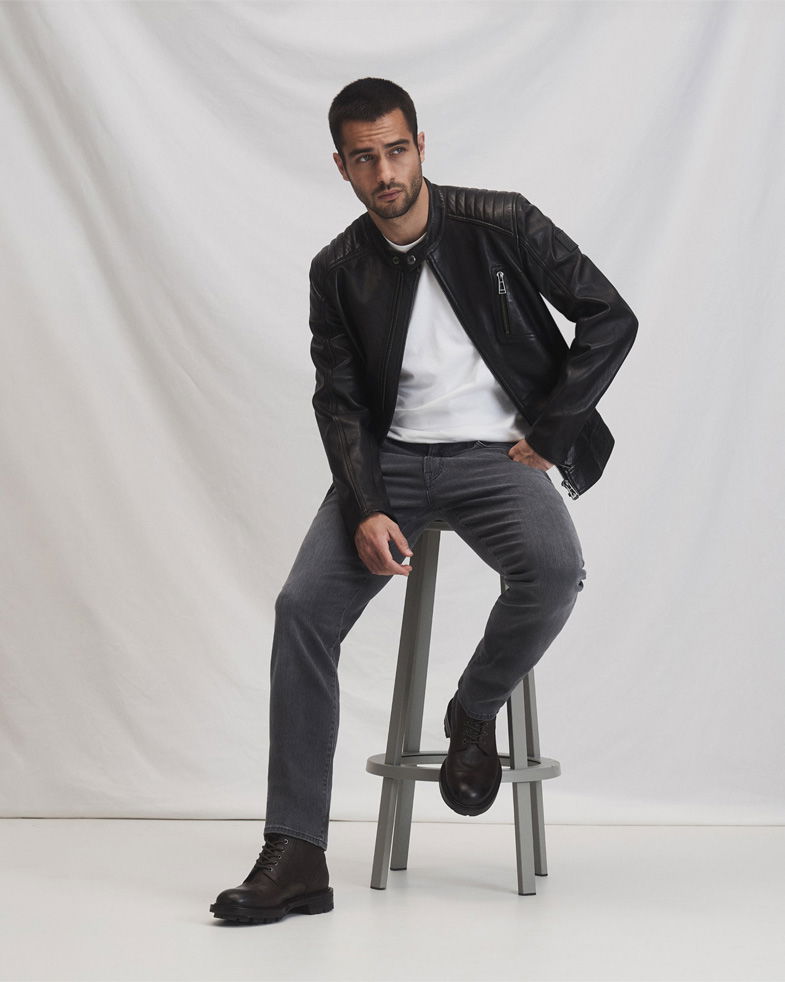 Mann sitzt auf grauem Hocker, trägt schwarze Lederjacke, weißes T-Shirt, graue Jeans und schwarze Schnürboots.