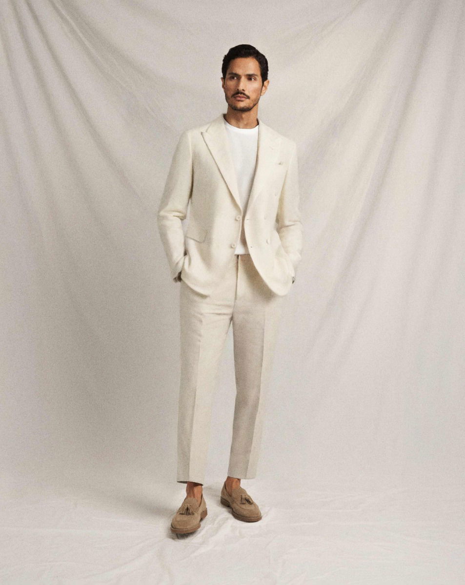 Mann steht vor hellem Hintergrund, trägt cremefarbenen Leinenanzug, weißes Shirt und hellbraune Tasselloafer Schuhe.