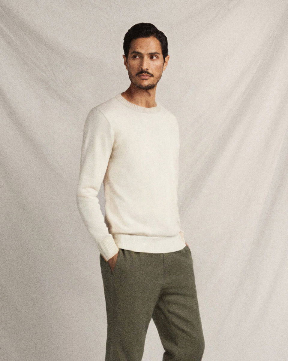 Mann mit dunklem Haar trägt cremefarbenen Rundhalspullover und olivgrüne Leinenhose, Hände in Taschen, vor hellem Hintergrund.