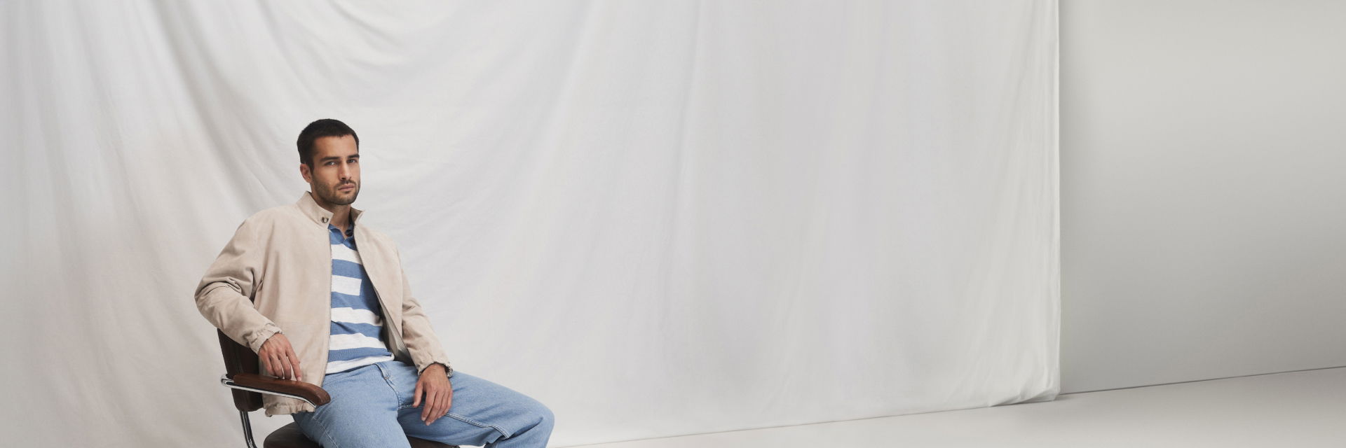 Mann sitzt auf Stuhl, trägt weiß-blau gestreiftes Poloshirt, hellblaue Jeans und beige Velourslederblouson vor weißem Hintergrund. Mann sitzt auf Stuhl, trägt weiß-blau gestreiftes Poloshirt, hellblaue Jeans und beige Velourslederblouson vor weißem Hintergrund.