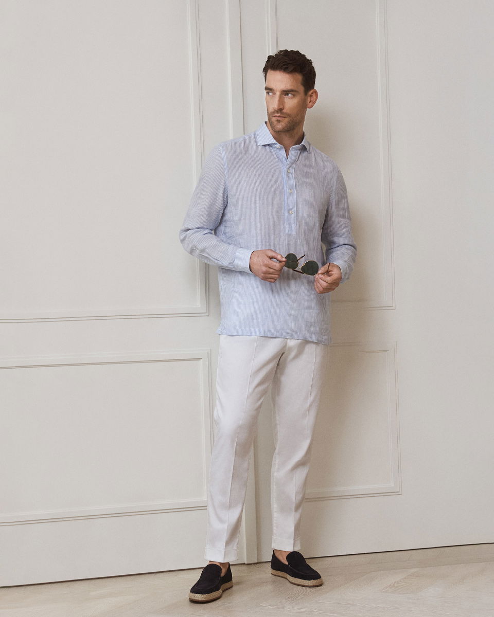 Mann trägt blau-weiß gestreiftes Popover-Hemd, weiße Hose, dunkelblaue Espadrilles und hält Sonnenbrille vor weißer Wand.