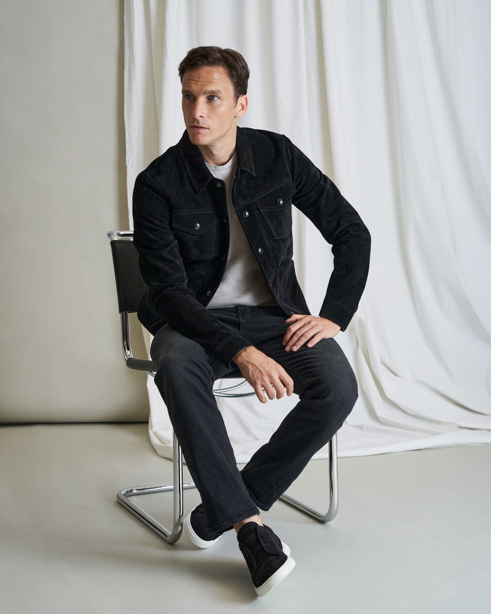 Mann sitzt auf Stuhl, trägt schwarzes Overshirt, grauen Pullover, schwarze Hose und schwarze Sneaker mit weißer Sohle.