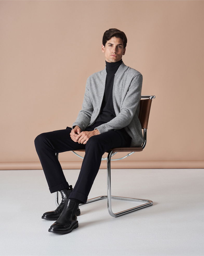 Mann sitzt auf Stuhl, trägt schwarzen Rollkragenpullover, schwarze Hose, hellgraues Blouson und schwarze Schnürboots. Mann sitzt auf Stuhl, trägt schwarzen Rollkragenpullover, schwarze Hose, hellgraues Blouson und schwarze Schnürboots.