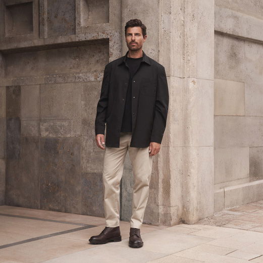 Mann trägt schwarzen Rundhalspullover, schwarzes Overshirt, beige Chinohose und braune Schnürboots, steht vor Steinmauer. Mann trägt schwarzen Rundhalspullover, schwarzes Overshirt, beige Chinohose und braune Schnürboots, steht vor Steinmauer.