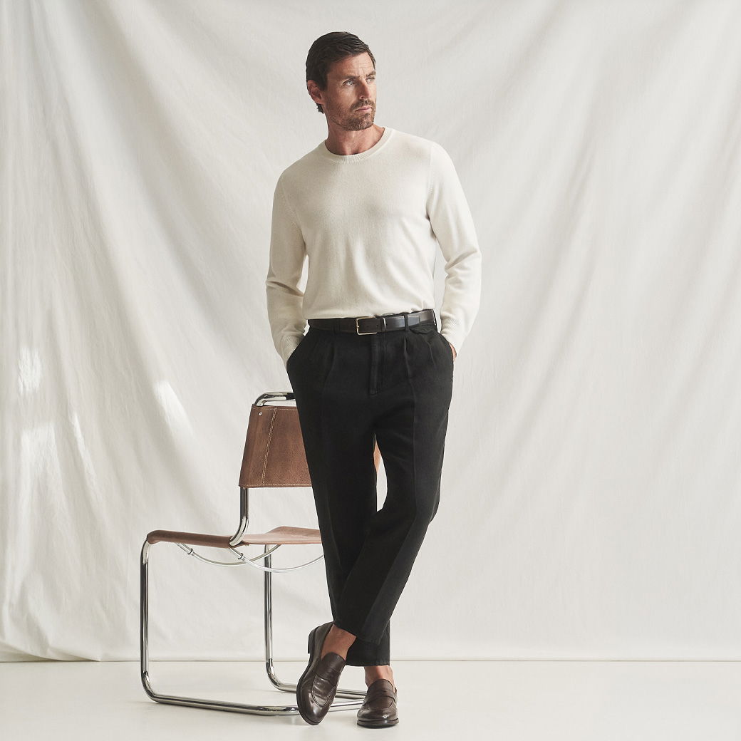 Mann steht vor weißem Hintergrund, trägt cremefarbenen Rundhalspullover, schwarze Hose, schwarzen Gürtel und braune Lederloafer.
