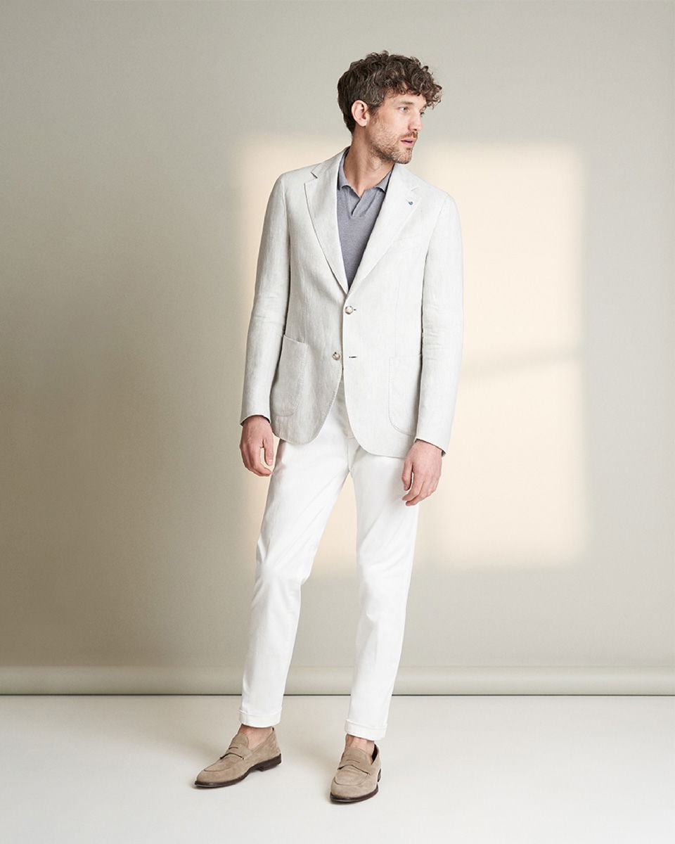 Mann trägt hellgraues Sakko, graues Strickpolo, weiße Hose und beige Loafer, steht vor hellem Hintergrund.