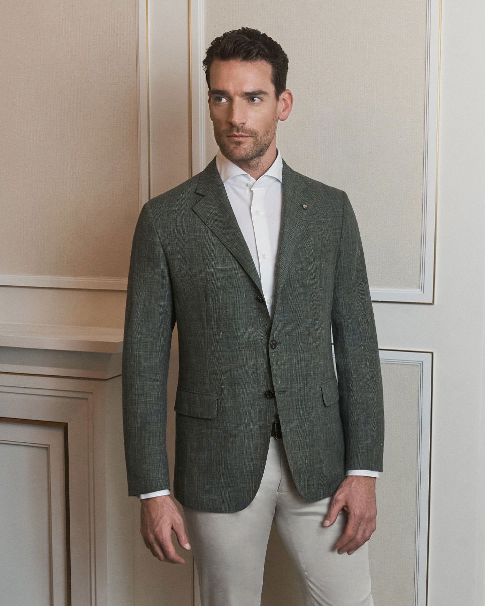 Mann steht vor Wand, trägt dunkelgraues Sakko, weißes Hemd und beigefarbene Chinohose, blickt seitlich weg.