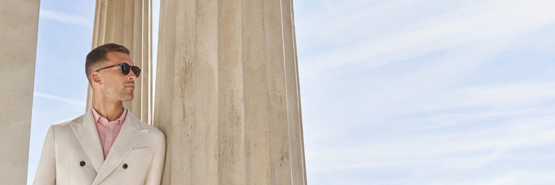 Mann mit cremefarbenem Sakko, rosa Hemd und schwarzer Sonnenbrille lehnt an Säule vor blauem Himmel. Mann mit cremefarbenem Sakko, rosa Hemd und schwarzer Sonnenbrille lehnt an Säule vor blauem Himmel.