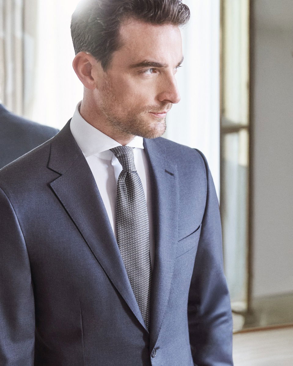 Mann mit gepflegtem Bart trägt navyblaues Sakko, weißes Hemd und silber-gemusterte Krawatte, Blick nach rechts.