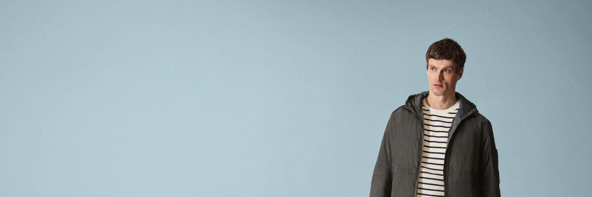 Mann mit kurz geschnittenem braunen Haar trägt blau-weiß gestreiftes Shirt und graue Kapuzenjacke vor hellblauem Hintergrund. Mann mit kurz geschnittenem braunen Haar trägt blau-weiß gestreiftes Shirt und graue Kapuzenjacke vor hellblauem Hintergrund.