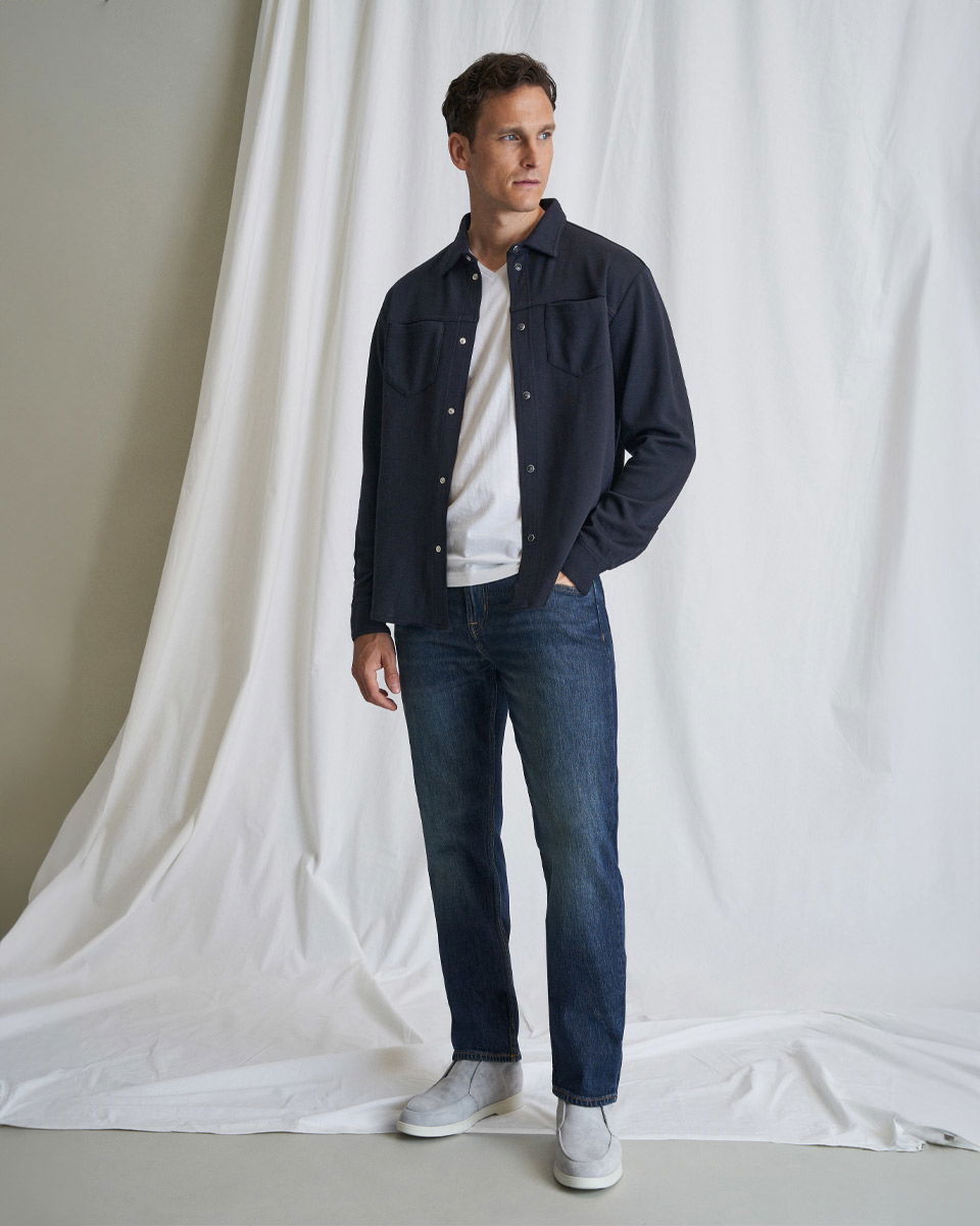 Mann trägt weißes T-Shirt, dunkelblaues Overshirt, dunkle Jeans und hellgraue Loafer vor weißem Hintergrund.