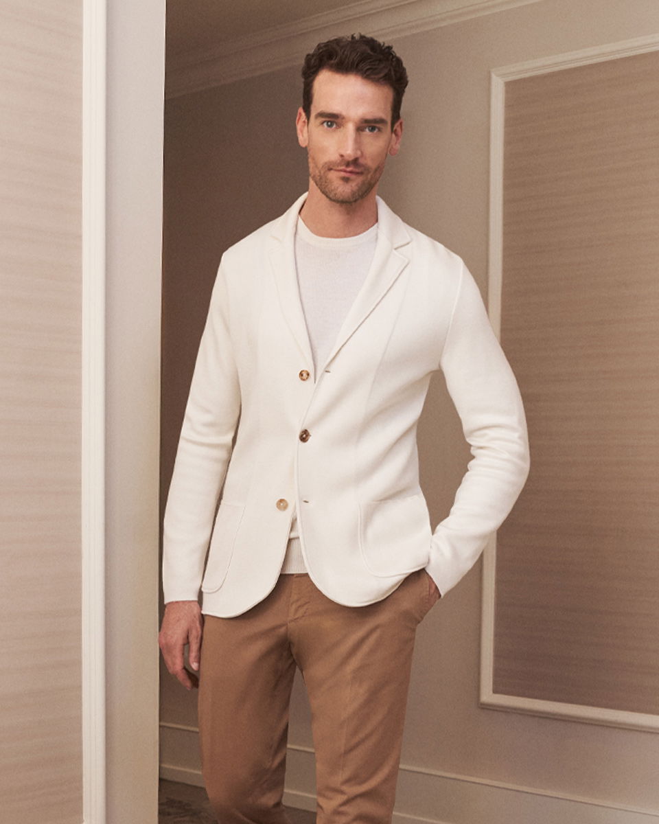 Mann mit weißem Sakko, weißem Shirt und brauner Chinohose steht lässig in elegantem Innenraum vor Wand mit Bilderrahmen.