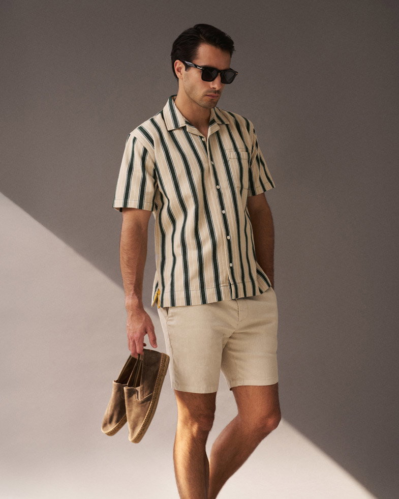 Mann mit beige-grün gestreiftem Kurzarmhemd, beigen Shorts und braunen Espadrilles in der Hand vor grauem Hintergrund. Mann mit beige-grün gestreiftem Kurzarmhemd, beigen Shorts und braunen Espadrilles in der Hand vor grauem Hintergrund.